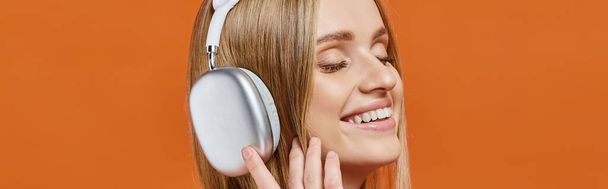 ritratto di donna bionda soddisfatta nell'ascoltare musica in cuffia con gli occhi chiusi su arancione, striscione - Foto, immagini