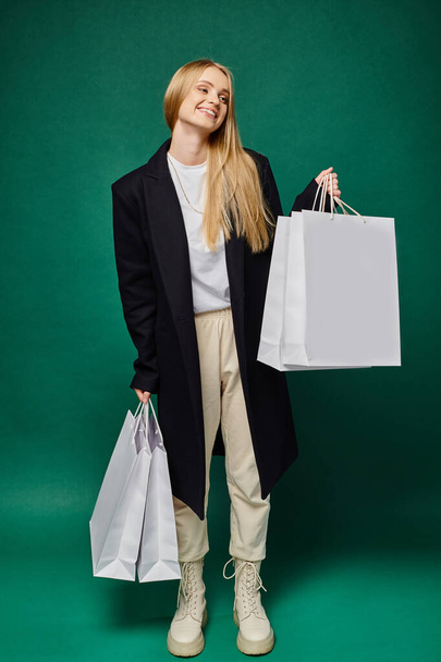 full length of joyful blonde woman in black coat holding white shopping bags on green, winter sales - Photo, Image