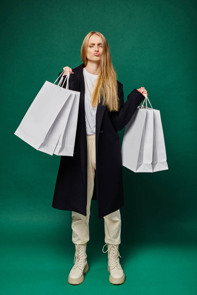 trendy blondýnka v černém kabátě drží bílé nákupní tašky a našpulené rty na zelené, plné délce - Fotografie, Obrázek