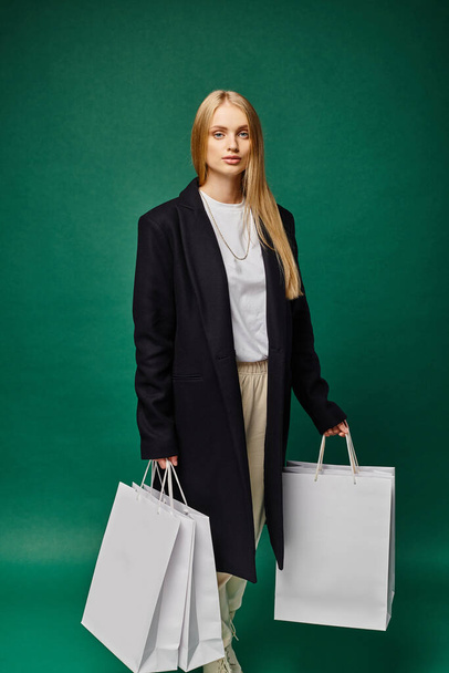 pewny blondynka kobieta w czarny płaszcz gospodarstwa białe torby na zakupy i patrząc na aparat na zielony - Zdjęcie, obraz