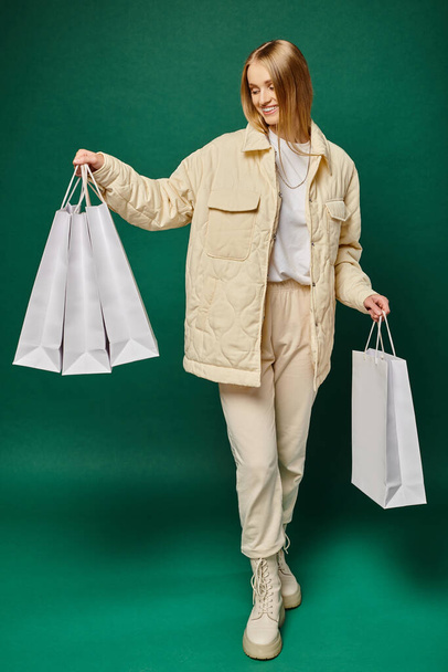 full length of cheerful blonde woman in trendy winter attire holding white shopping bags on green - Photo, Image
