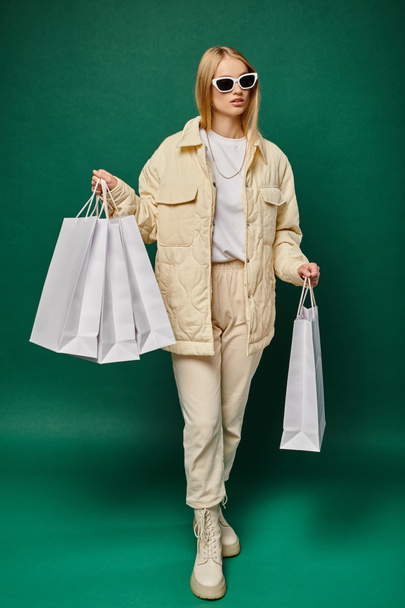 stylish blonde woman in winter wear and sunglasses walking with shopping bags on green, full length - Photo, Image