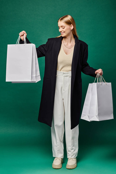 full length of pleased blonde woman in black warm coat standing with white shopping  bags on green - Photo, Image