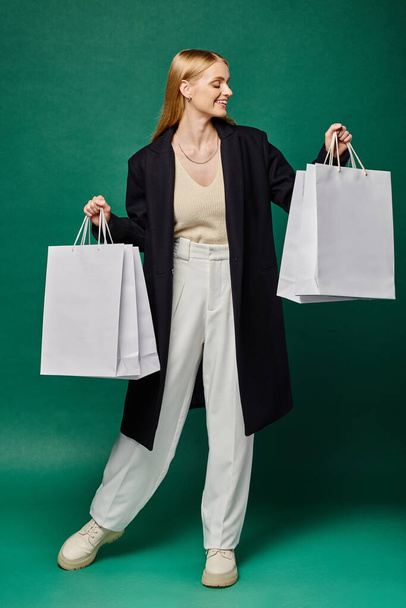 delighted blonde woman in black stylish coat with white shopping  bags on green, winter sales - Photo, Image