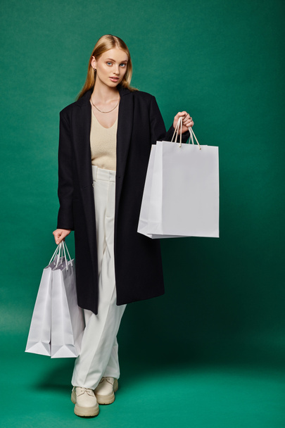 volledige lengte van stijlvolle blonde vrouw in zwarte jas poseren met witte boodschappentassen op groen - Foto, afbeelding