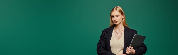 fashionable blonde woman in black winter coat standing with laptop on green backdrop, banner - Photo, Image