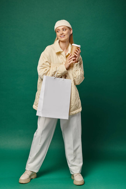 young smiling woman in trendy winter clothing with shopping bag and coffee to go on green backdrop - Photo, Image