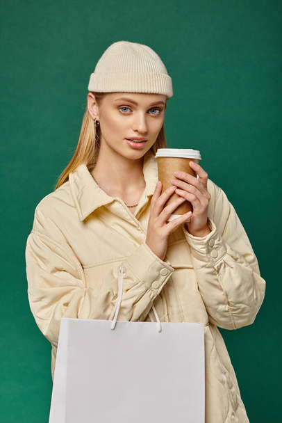 очаровательная женщина в теплой куртке и шляпе шапочка с мешком для покупок и бумажной чашкой на зеленый, зимний стиль - Фото, изображение