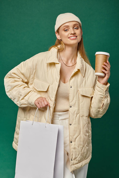 joyful woman in warm winter clothes with shopping bag and paper cup looking at camera on green - Photo, Image