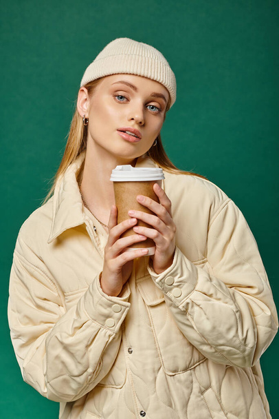 charming woman in beanie hat and warm stylish jacket with coffee to go on green, winter emotions - Photo, Image