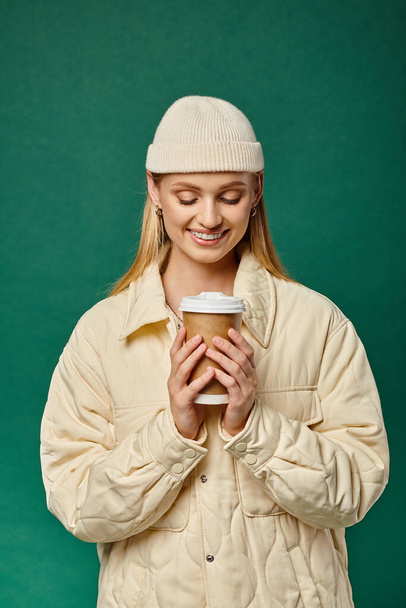 zadowolona kobieta w czapce i ciepłej modnej kurtce trzyma gorący napój na wynos na zielono, zimowa radość - Zdjęcie, obraz
