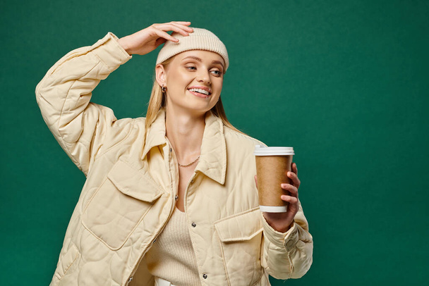 mujer alegre en gorro sombrero y chaqueta caliente sosteniendo bebida caliente para llevar y mirando hacia otro lado en verde - Foto, imagen