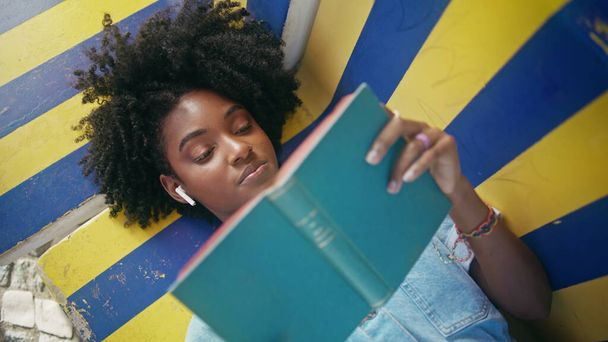Lähikuva teini käsittelyssä kirja ääneen makaa värikäs penkki ulkona. Curly afrikkalainen amerikkalainen tyttö kuulokkeet nauttia kirjallisuudesta harrastus. Kaunis nuori nainen rentouttava suosikki romaani kesällä viikonloppuna - Valokuva, kuva