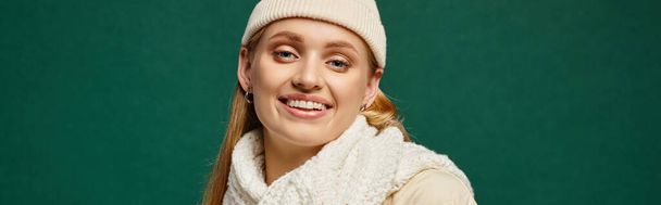 trendy woman in white knitted scarf and beanie hat smiling at camera on green, winter style banner - Photo, Image