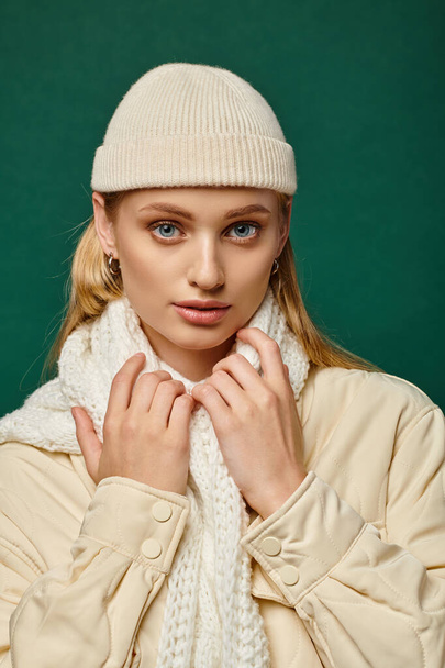 young woman in warm jacket and beanie hat with white scarf looking at camera on green, winter trend - Foto, imagen