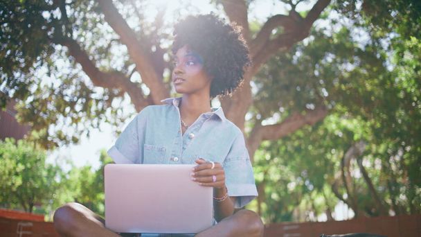 Kudrnatý teenager sedí s laptopem na slunci a rozhlíží se. Krásná africká americká dospívající dívka drží notebook relaxaci na slunné přírodě. Bezstarostná mladá žena odpočívat po vzdálené práci venku. - Fotografie, Obrázek