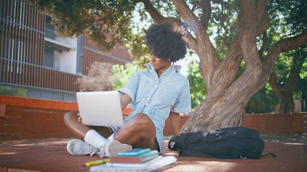 Afrykański student wpisując laptop siedzi słoneczny park w bezprzewodowych słuchawkach. Cute czarnowłosa dziewczyna zmęczona studiami online na letniej przyrody. Modny atrakcyjny nastolatek patrząc komputer pod zielonym drzewem. - Zdjęcie, obraz