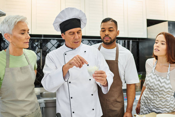 gut aussehende reife Koch diskutiert mit seinen multirassischen Studenten, wie man backt, Süßwaren - Foto, Bild