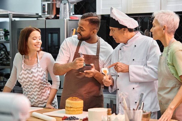 Веселая молодая женщина смотрит на своих многорасовых друзей и шеф-повара, студентка чистит торт сиропом - Фото, изображение