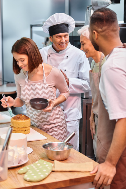 giovane donna allegra spazzolatura torta con sciroppo mentre i suoi diversi amici e chef parlando attivamente - Foto, immagini