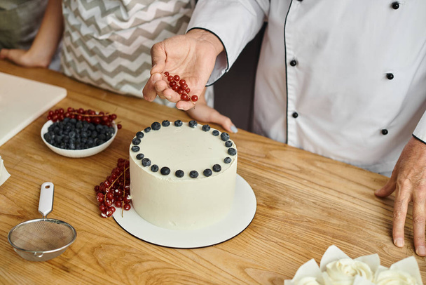 vue recadrée du chef mature décorant délicieux gâteau avec groseille rouge à côté de son étudiante - Photo, image