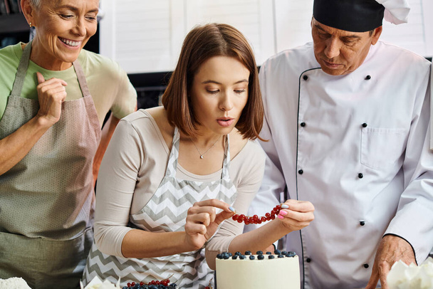 junge Frau mit Schürze dekoriert Kuchen mit roter Johannisbeere neben ihrem fröhlichen reifen Freund und Koch - Foto, Bild
