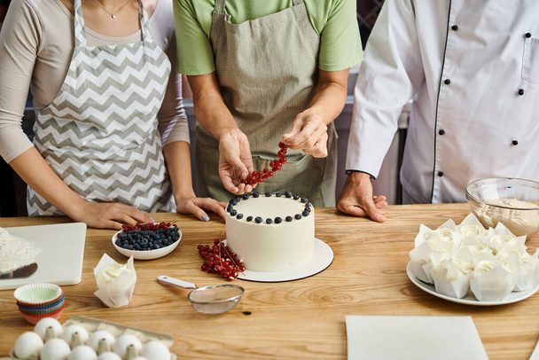 обрізаний вид зрілої жінки, що прикрашає торт з ягодами поруч з шеф-кухарем та її молодим другом - Фото, зображення
