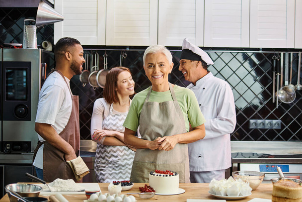 Сосредоточьтесь на зрелой женщине с тортом улыбаясь в камеру с размытыми разнообразными друзьями и шеф-поваром на заднем плане - Фото, изображение