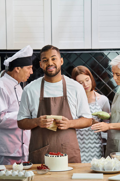 focus op vrolijke Afrikaanse Amerikaanse man met taart glimlachen op de camera met zijn wazige vrienden op de achtergrond - Foto, afbeelding