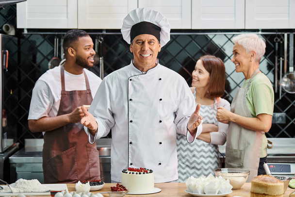 se centran en alegre cocinero maduro posando con pastel y sonriendo a la cámara, sus estudiantes borrosos en el fondo - Foto, imagen