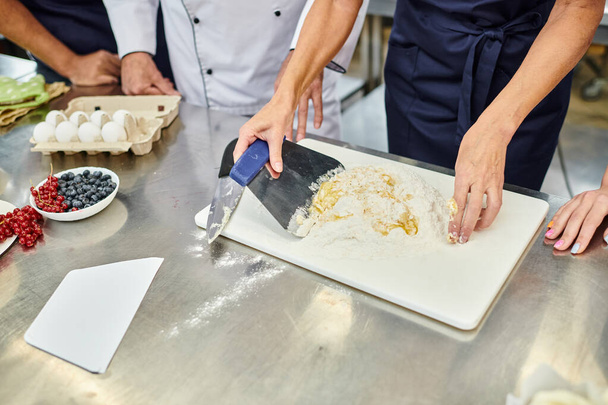 cropped view of mature female chef working with dough next to her colleagues, confectionary - Fotografie, Obrázek