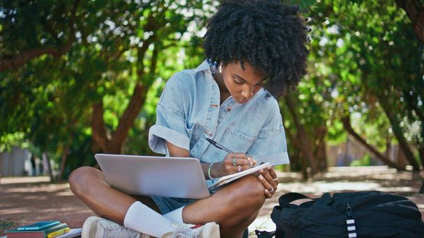 Tyttö kannettavan tehdä muistiinpanoja muistilehtiössä istuu vihreä luonto lähikuva. Keskittynyt afrikkalainen amerikkalainen naisopiskelija opiskelee verkossa kirjoittamista muistikirjassa kadulla. Curly kiireinen nainen oppiminen kurssi ulkona - Valokuva, kuva