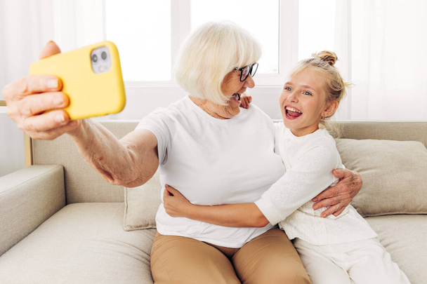 Isoäiti lapsi selfie sohva liimaus t-paita kopioi onnea yhdessä video puhelin koulutus valokuvaus halaus perhe ihmiset kaksi tilaa sisätiloissa pojantytär soittaa valkoinen hymyillen - Valokuva, kuva