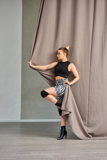 mulher golpeia uma pose em um movimento de dança gracioso, equilibrando em um pé contra uma parede coberta de cortina - Foto, Imagem