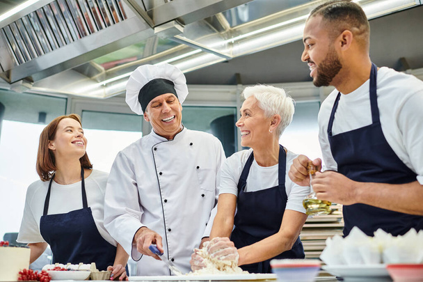 gioioso capo cuoco maturo in cappello bianco aiutare i suoi chef multiculturali con pasta, pasticceria - Foto, immagini