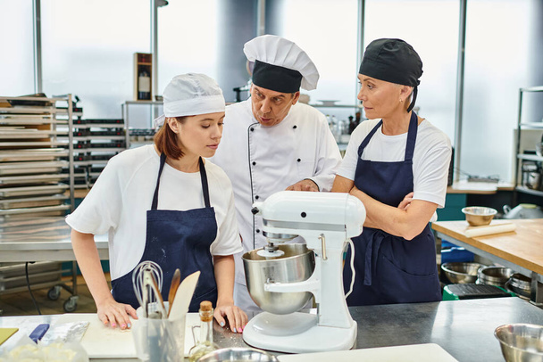 knappe volwassen chef-kok uit te leggen hoe te werken met mixer aan zijn vrouwelijke chef-koks, zoetwaren - Foto, afbeelding