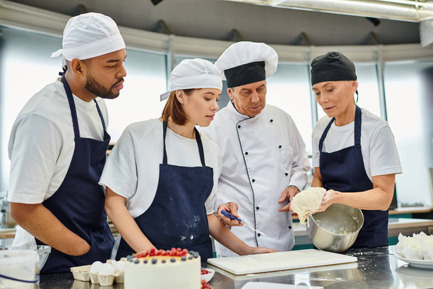 guapo cocinero jefe maduro en sombrero blanco viendo cómo sus diversos chefs en delantales trabajando con masa - Foto, imagen