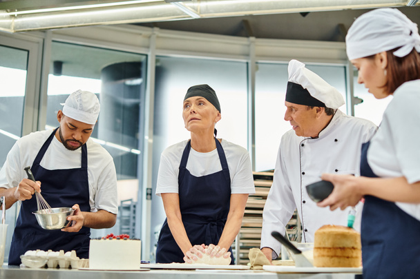 goed uitziende volwassen chef-kok praten met zijn hard werkende interraciale chefs terwijl op keuken - Foto, afbeelding