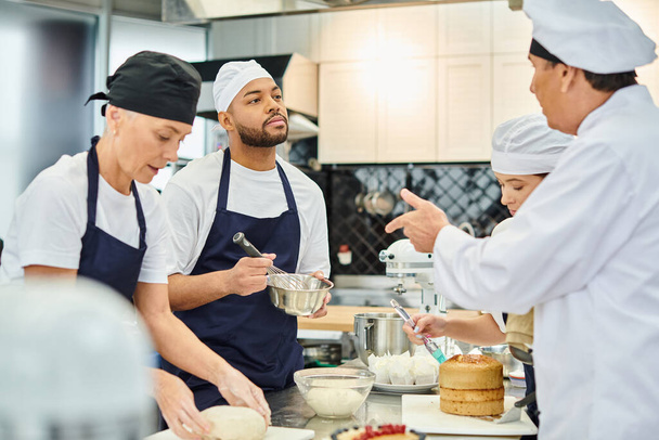 cozinheiro chefe maduro conversando com o jovem chef afro-americano enquanto seus colegas trabalham em pastelaria - Foto, Imagem