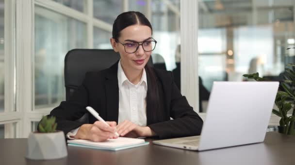 Skupiona bizneswoman nosząca okulary siedząca przy stole i pracująca z nowoczesnym laptopem podczas pisania informacji w notebooku w jasnym biurze. Pojęcie samorozwoju i odpowiedzialności. - Materiał filmowy, wideo