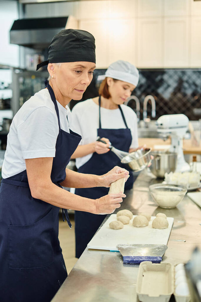 focus op volwassen chef-kok in toque en schort werken met deeg naast haar jonge wazig collega - Foto, afbeelding