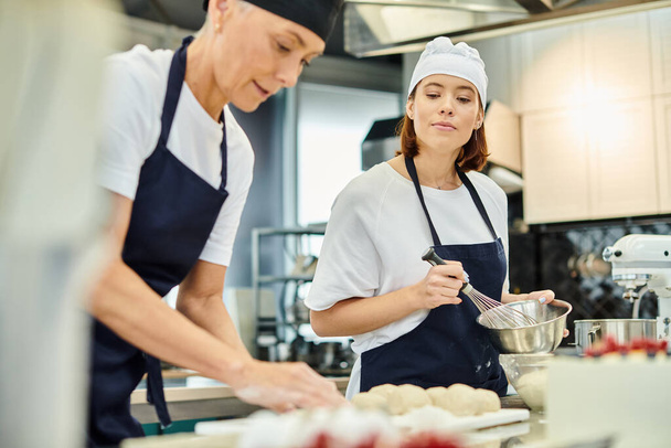 aantrekkelijke jonge chef-kok in schort kijken naar haar volwassen collega werken aan deeg, zoetwaren - Foto, afbeelding