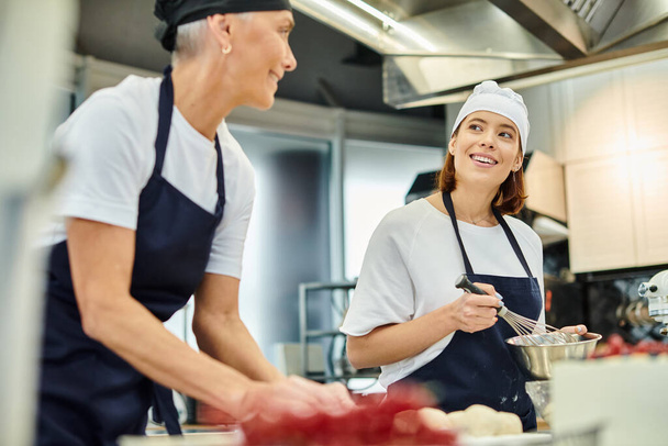 centrarse en la alegre joven chef en delantal sonriendo a su borrosa colega madura mientras trabaja en la pastelería - Foto, imagen