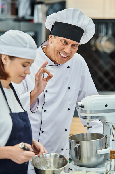 Сосредоточьтесь на зрелом веселом шеф-поваре в белой шляпе, показывая нормальный жест рядом с его размытой женщиной-поваром - Фото, изображение