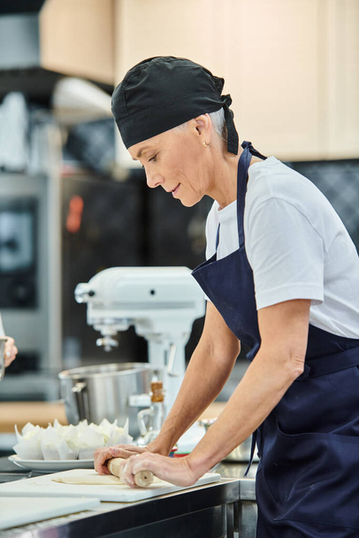 зрелая веселая женщина шеф-повар в синем токе и фартуке работает с тестом на кухне, кондитерские изделия - Фото, изображение