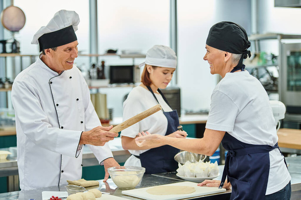 beau chef cuisinier joyeux donnant rouleau à pâtisserie à son chef féminin à côté de leur jeune collègue - Photo, image