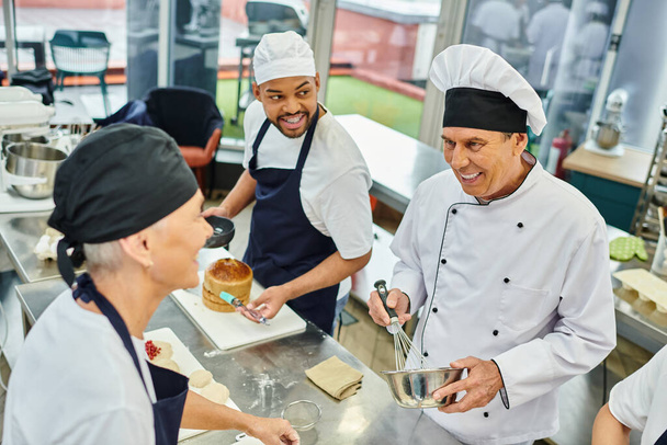 весела багаторасова команда на шеф-кухарях в синіх тонах дивиться на їх зрілий головний кухар, кондитерські вироби - Фото, зображення