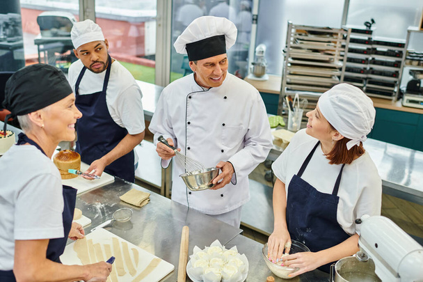 guapo jefe de cocina alegre en sombrero blanco rodeado de sus chefs multirraciales, confitería - Foto, imagen