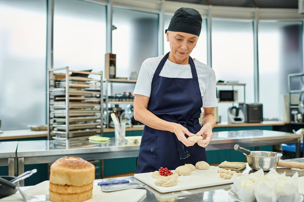 привлекательная зрелая женщина в синем фартуке и токе, работающая с тестом на кухне, кондитерских изделиях - Фото, изображение