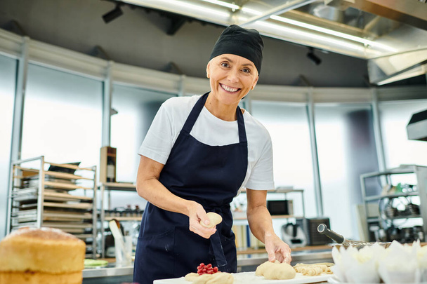 atractivo cocinero maduro en toque azul sonriendo a la cámara mientras trabaja con masa, confitería - Foto, imagen